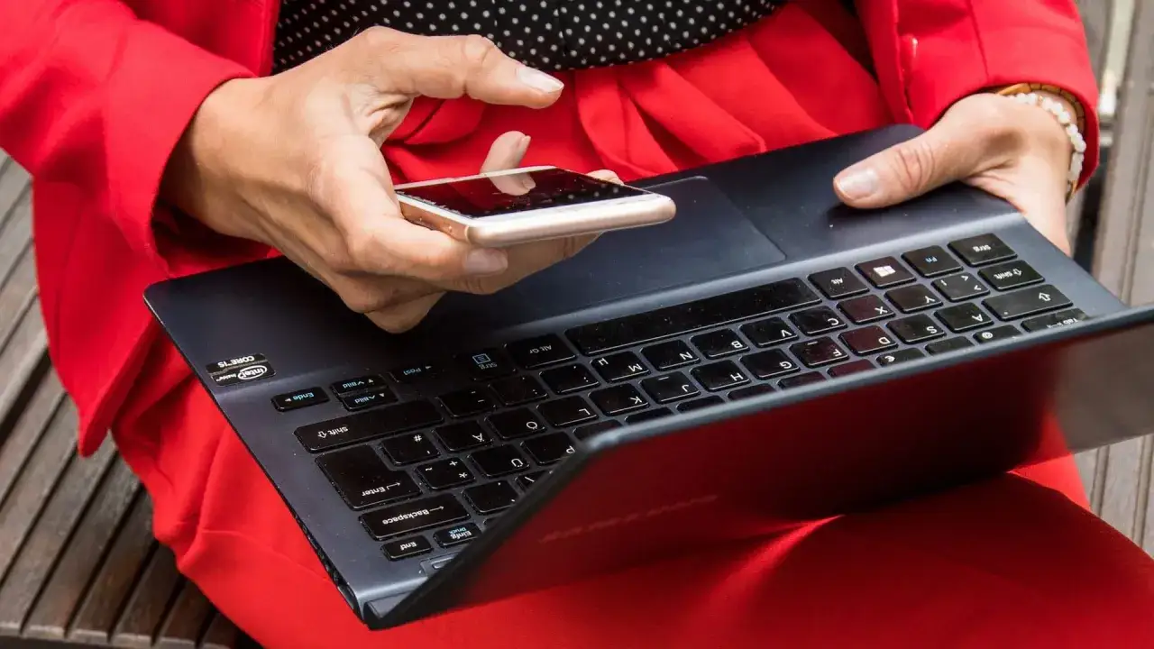 Besorgte Person prüft mit dem Handy, ob die mailadresse gehackt wurde. Roter Blazer, Laptop.