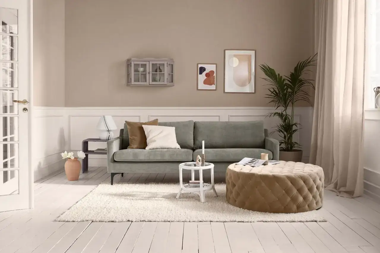 Gem&uuml;tliches Wohnzimmer mit grauem Sofa, beigem Pouf und einem Hauch von mauve farbe an der Wand.