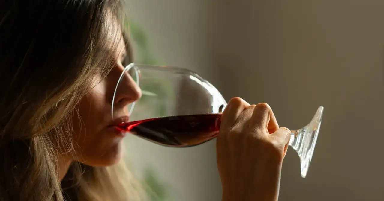 Kobieta pije czerwone wino z kieliszka. Ciemne włosy opadają na jej twarz, gdy unosi szkło do ust.