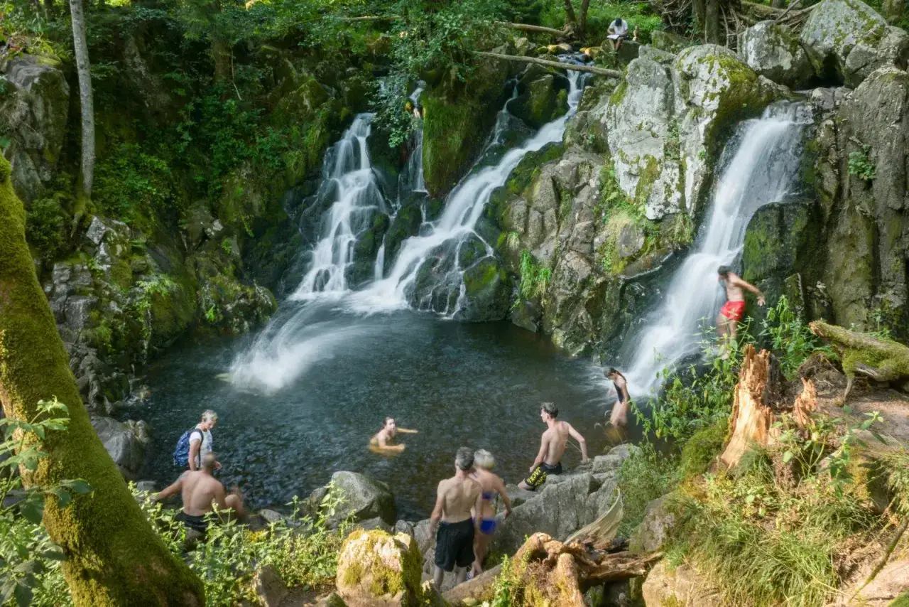 Des baigneurs profitent d'un saut du bouchot dans une cascade rocheuse entour&eacute;e de verdure.