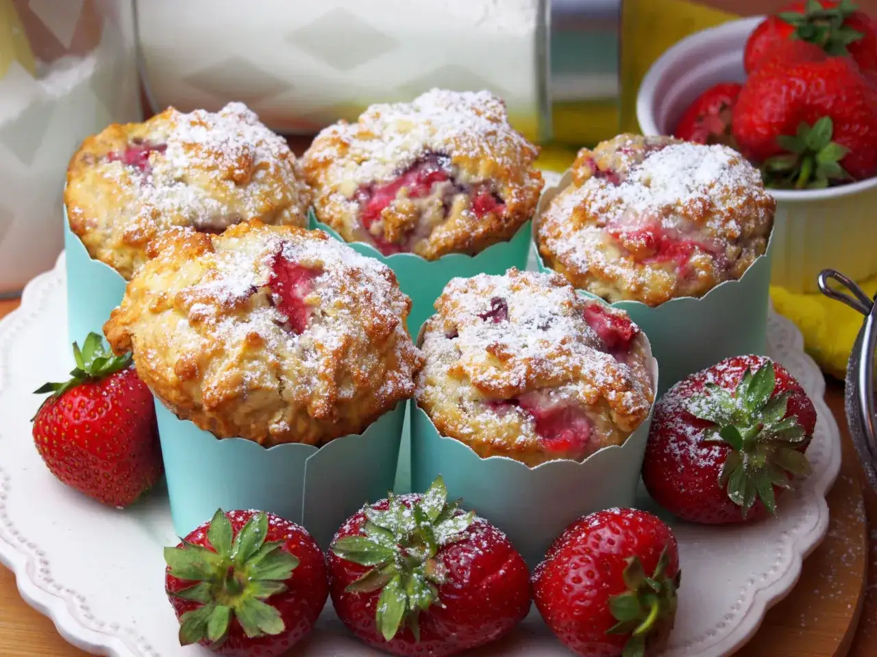 Pyszne muffiny z truskawkami, posypane cukrem pudrem, prezentują się apetycznie obok świeżych owoców.