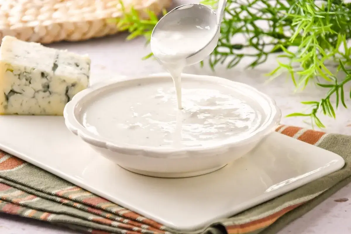 Salsa roquefort: cremosa y deliciosa, lista para servir. Un trozo de queso azul y una cuchara vierten la salsa en un cuenco.