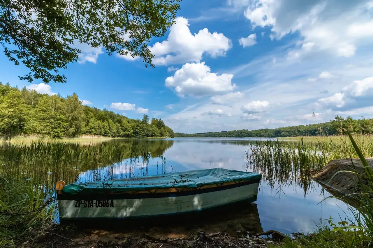 Ł&oacute;dka przy brzegu jeziora w ińskim parku krajobrazowym. Spokojna tafla wody odbija chmury i zielone drzewa.
