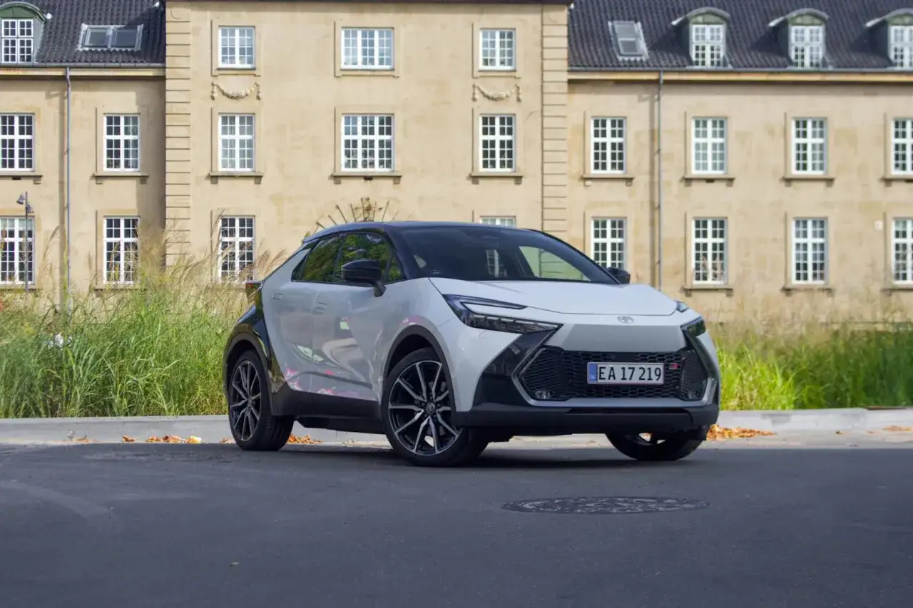 En vit Toyota C-HR Hybrid st&aring;r parkerad framf&ouml;r en gul byggnad med m&aring;nga f&ouml;nster. Bilen &auml;r redo f&ouml;r ett test.