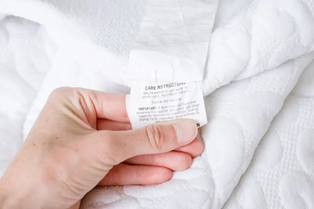 Hand holding a care tag with instructions on how to clean a mattress pad: machine wash cold, tumble dry low.