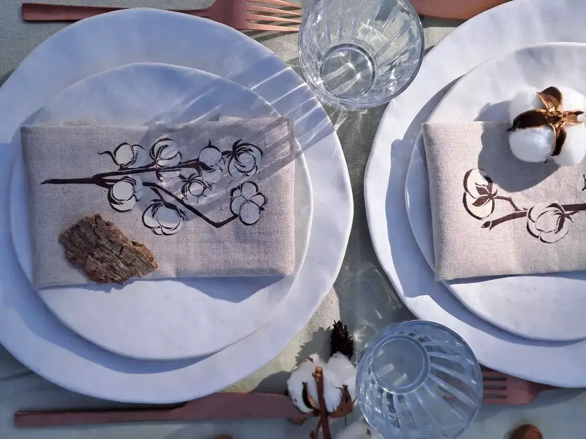 Table dress&eacute;e pour 1 an de mariage, noce de coton. Serviettes orn&eacute;es de branches de coton, assiettes blanches, verres textur&eacute;s et couverts cuivr&eacute;s.