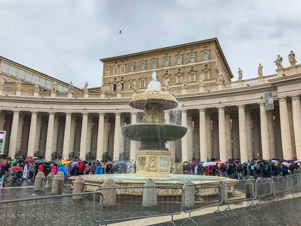 Plac Świętego Piotra, gdzie mieszka papież, z fontanną i kolumnadą Berniniego. Tłumy turystów z parasolami w deszczowy dzień.