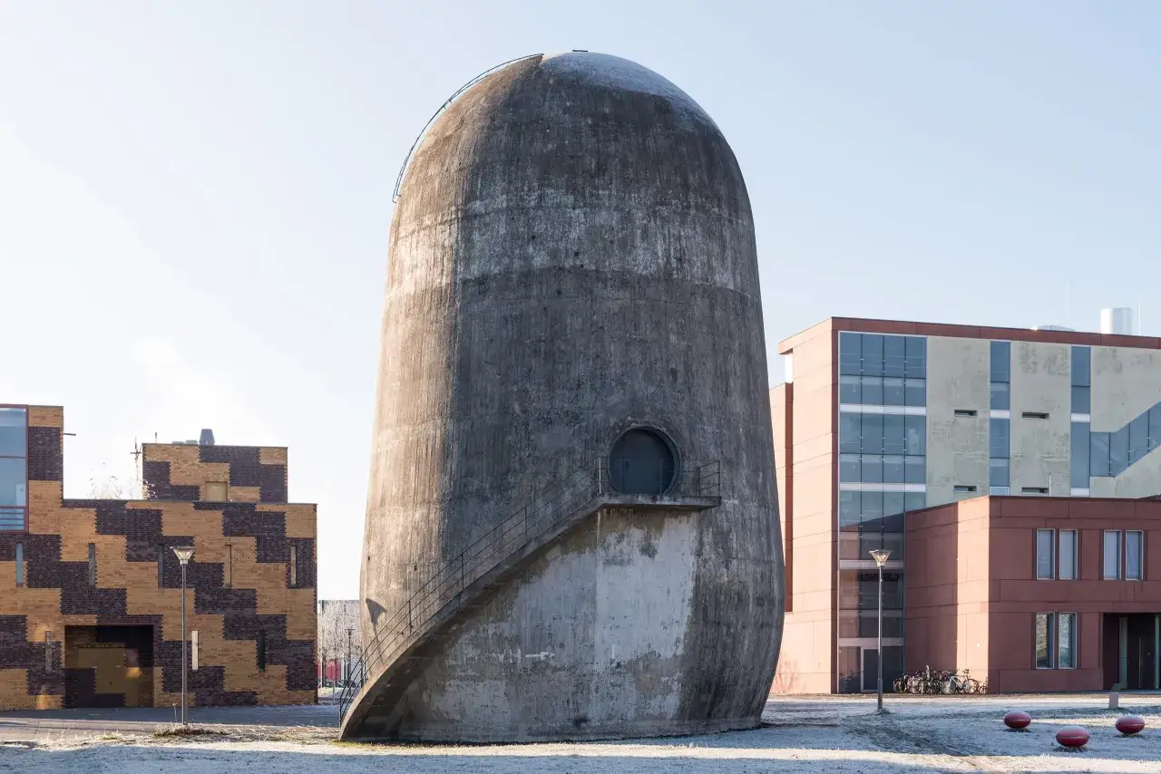 Der Trudelturm Berlin, ein markantes Betonbauwerk, ragt neben modernen Geb&auml;uden auf. Eine Au&szlig;entreppe f&uuml;hrt zu einer runden T&uuml;r.