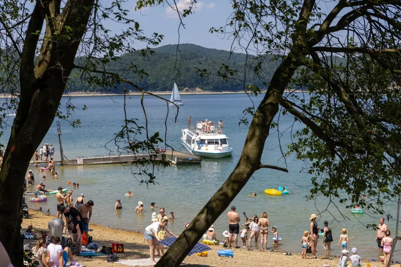 Polańczyk plaża tętni życiem. Ludzie kąpią się, opalają, a na wodzie pływa statek wycieczkowy i żagl&oacute;wka.
