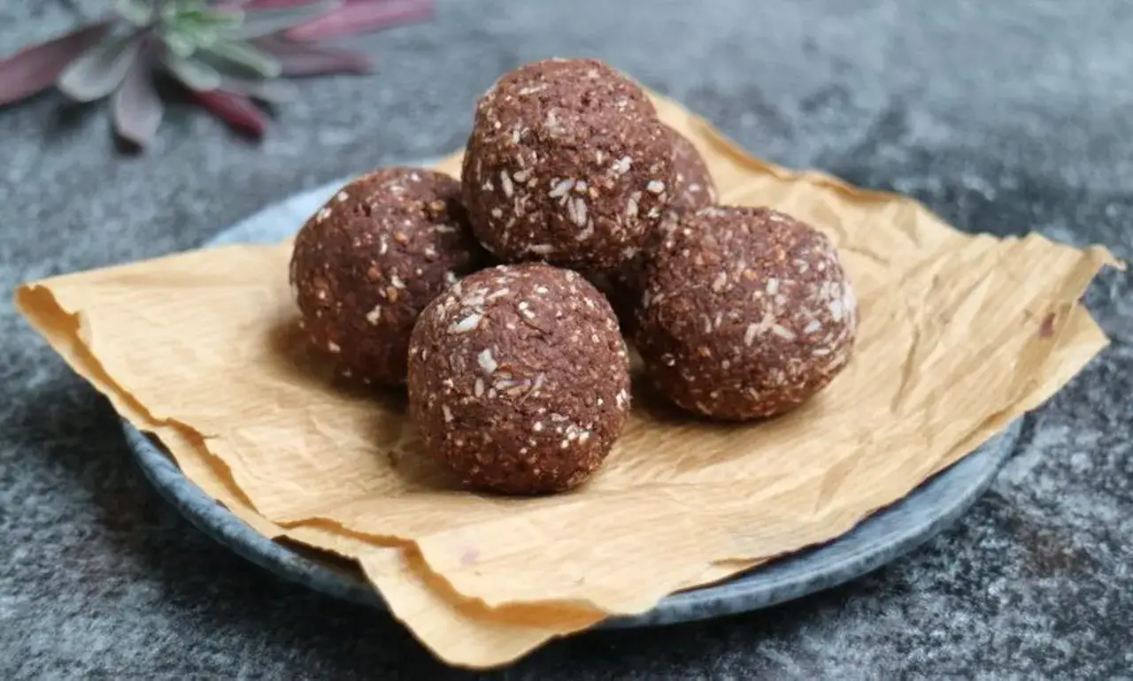 Fem nyttiga chokladbollar, rullade i kokos, ligger på ett fat med brunt papper. Perfekta för fika!
