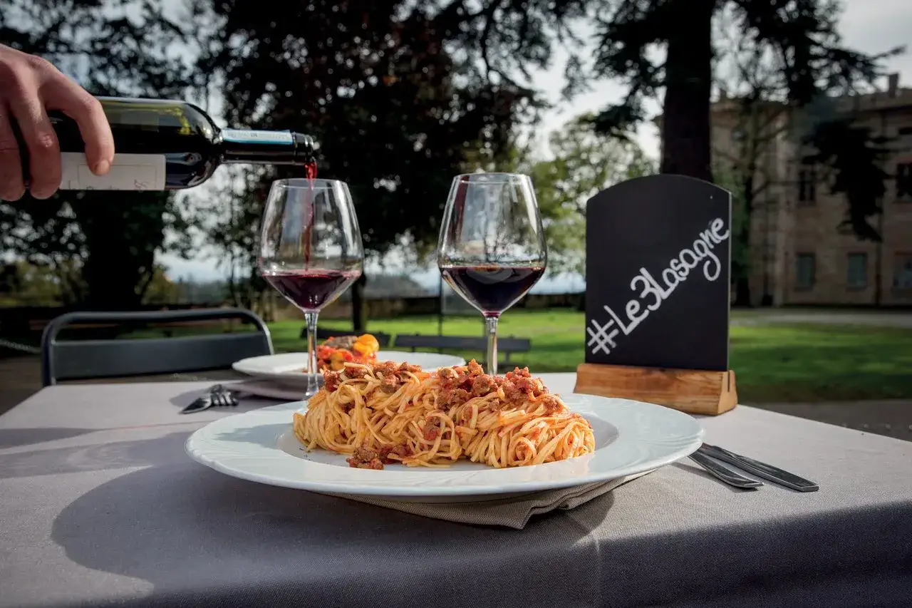 Rotwein wird in ein Glas gegossen, während eine Portion Spaghetti Bolognese auf einem Tisch im Freien steht. Die perfekte Antwort auf die Frage: welcher Wein zu Lasagne.