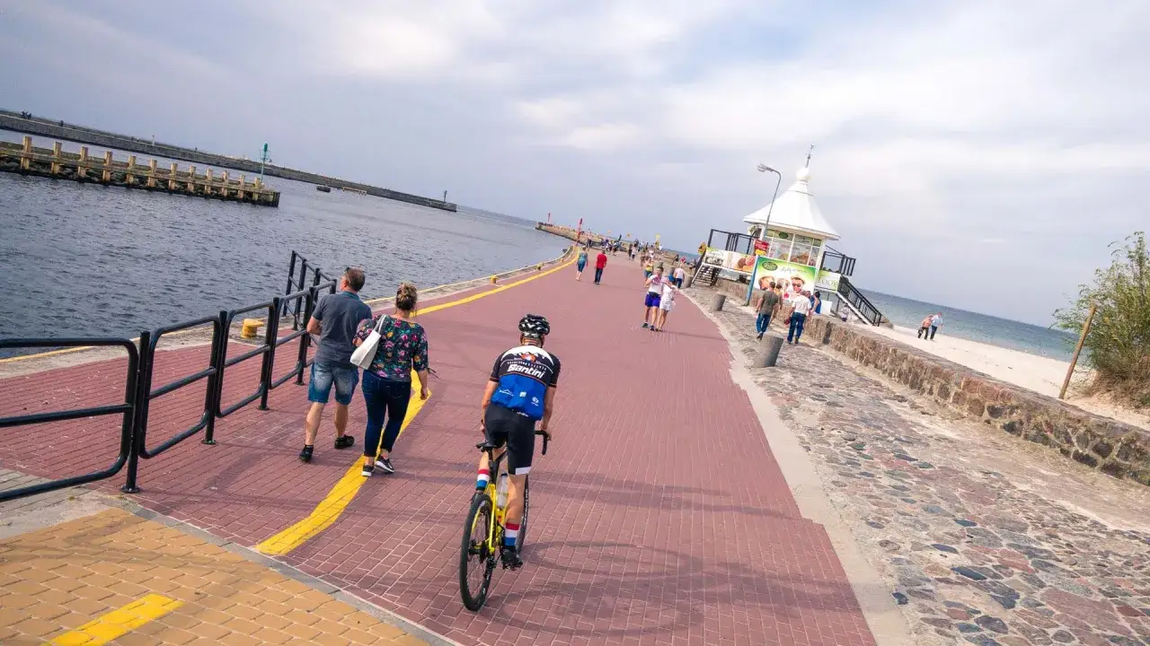Rowerzysta i spacerowicze na promenadzie w Słupsku. Trasa wzdłuż morza z widokiem na falochron i plażę.