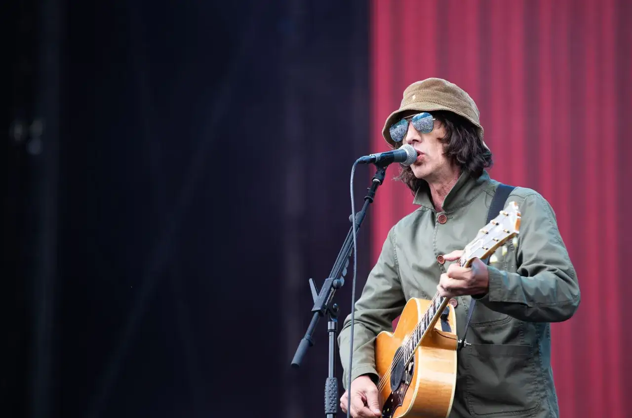 Richard Ashcroft, chanteur de The Verve, joue de la guitare et chante sur scène, portant un chapeau et des lunettes de soleil.