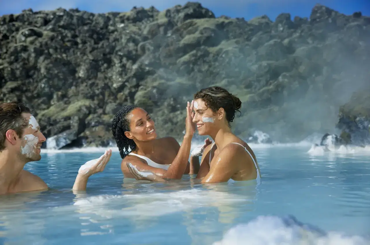 Trzy osoby w gorących źródłach na Islandii, nakładające sobie maski. Sprawdź blue lagoon islandia cena i zaplanuj relaks!