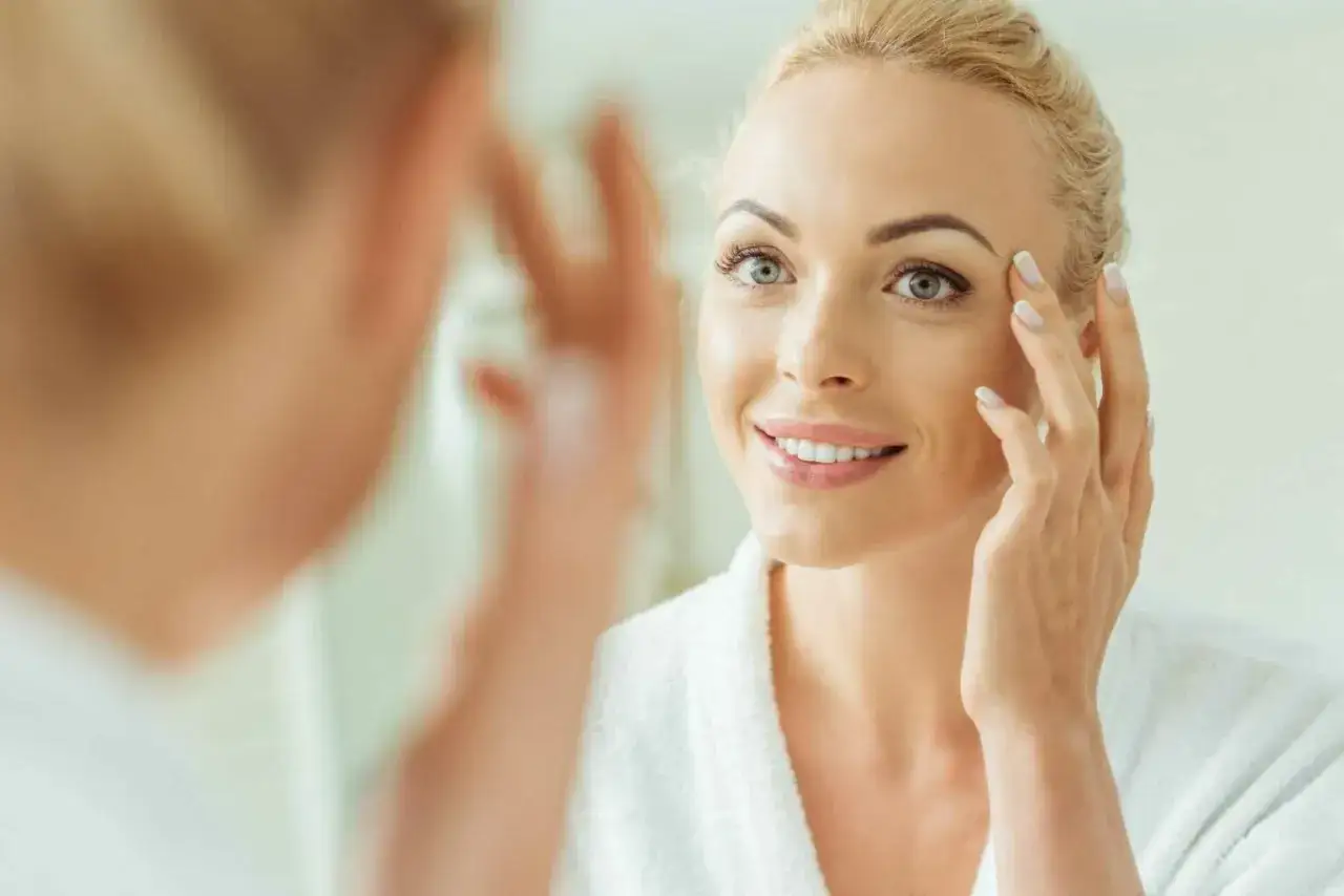 Une femme blonde souriante examine son visage dans un miroir, sa main effleurant sa joue. Elle pourrait utiliser un r&eacute;tino&iuml;de m&eacute;dicament pour une peau &eacute;clatante.