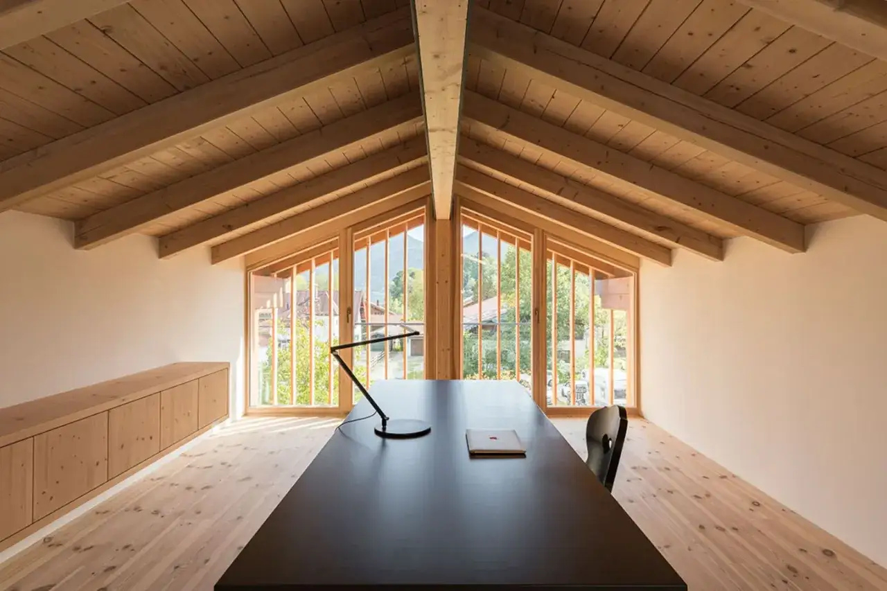 Ein moderner, offener Dachstuhl mit Holzbalken und gro&szlig;en Fenstern, die den Blick auf eine gr&uuml;ne Landschaft freigeben. Ein Schreibtisch mit Lampe und Stuhl steht im Raum.