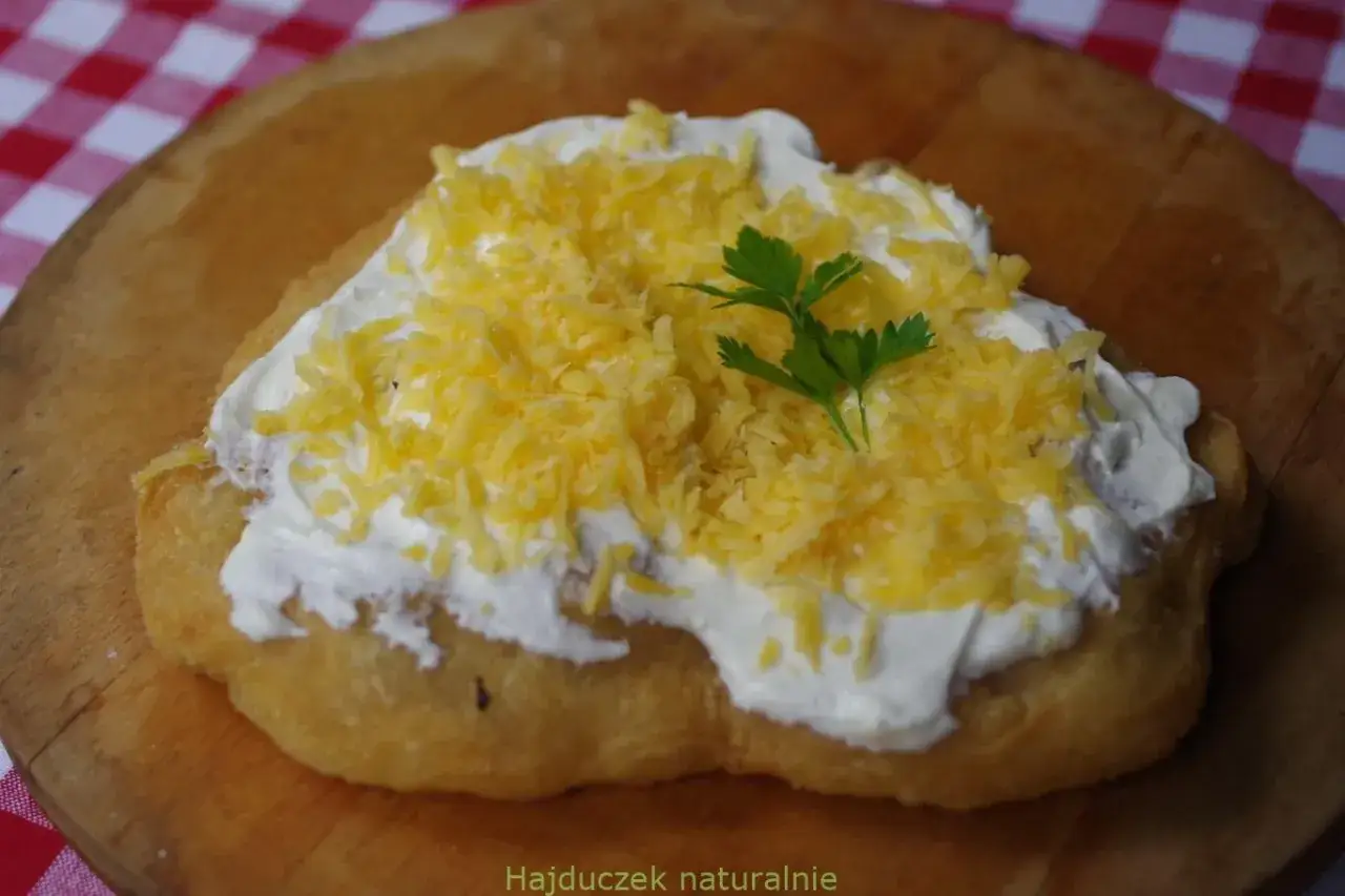 Lángos, tradycyjne danie kuchni węgierskiej, z obfitą porcją śmietany i tartego sera, ozdobiony natką pietruszki.