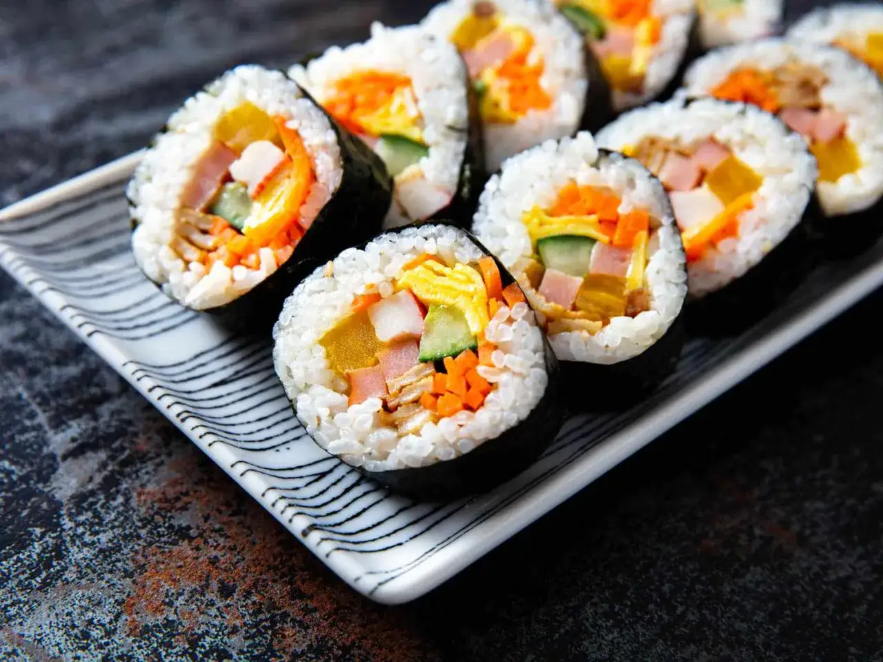 Sushi-Rollen mit Karotten, Gurken, Ei und Schinken. Was ist um Sushi herum? Eine gestreifte Platte und ein dunkler Hintergrund.