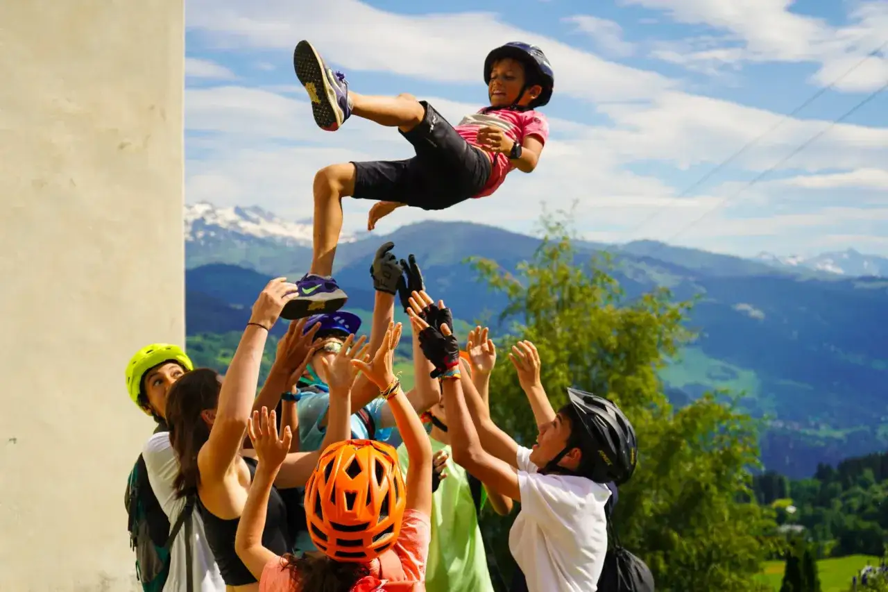 Kinder werden beim **Summer Camp** in die Luft geworfen. Ein Junge fliegt hoch, w&auml;hrend andere ihn auffangen. Berge im Hintergrund.