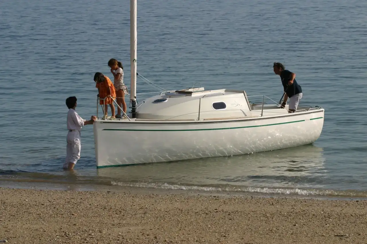 Une famille prépare leur voilier, icône 6, pour une sortie en mer. L'eau calme reflète le ciel bleu.