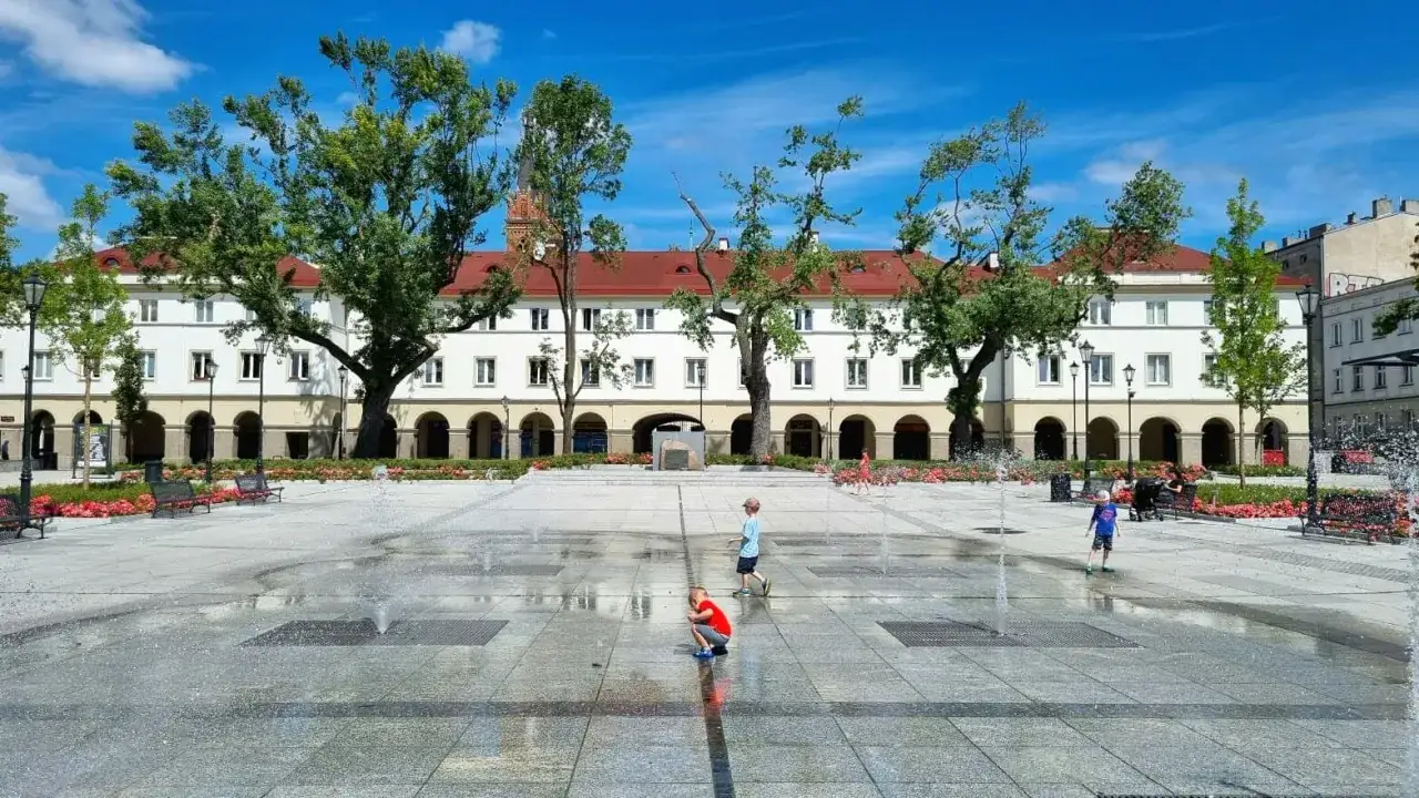 Dzieci bawią się na wodnej fontannie na łódzkim rynku. Słoneczny dzień, białe kamienice i zielone drzewa tworzą letnią atmosferę.