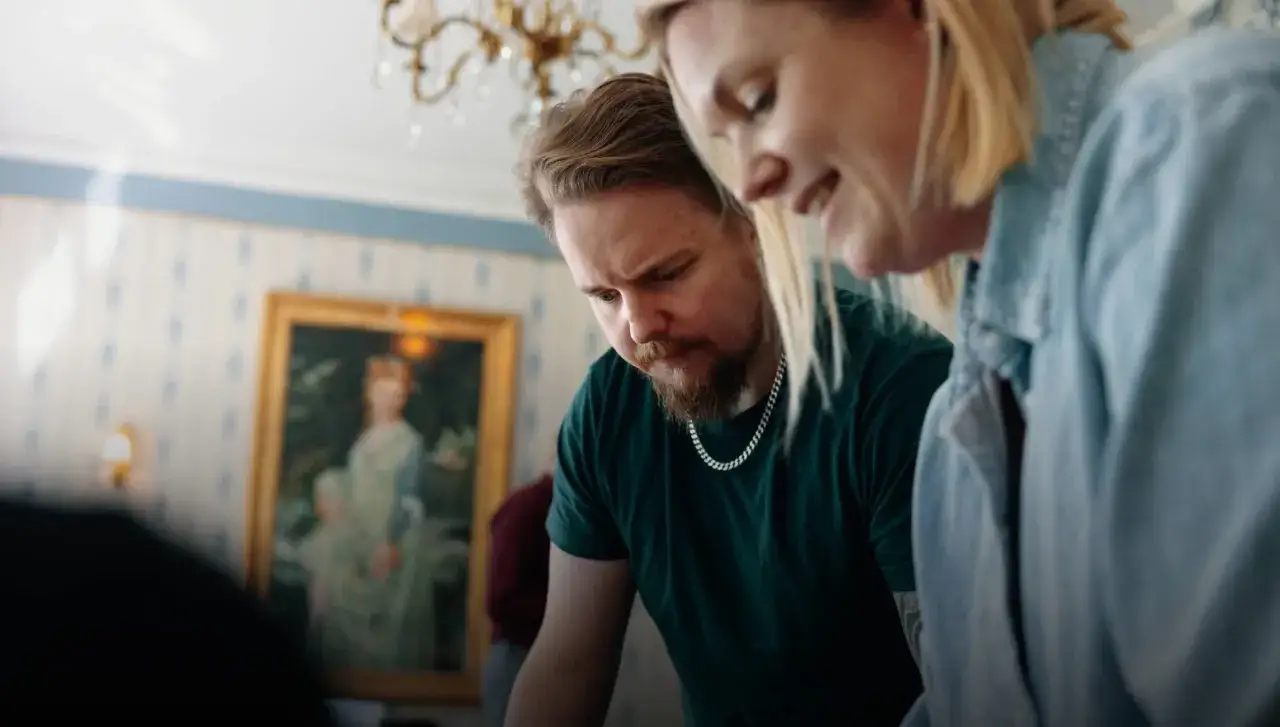 Två kollegor ler under en aktivitet med jobbet. I bakgrunden syns en tavla med en kvinna och ett barn.