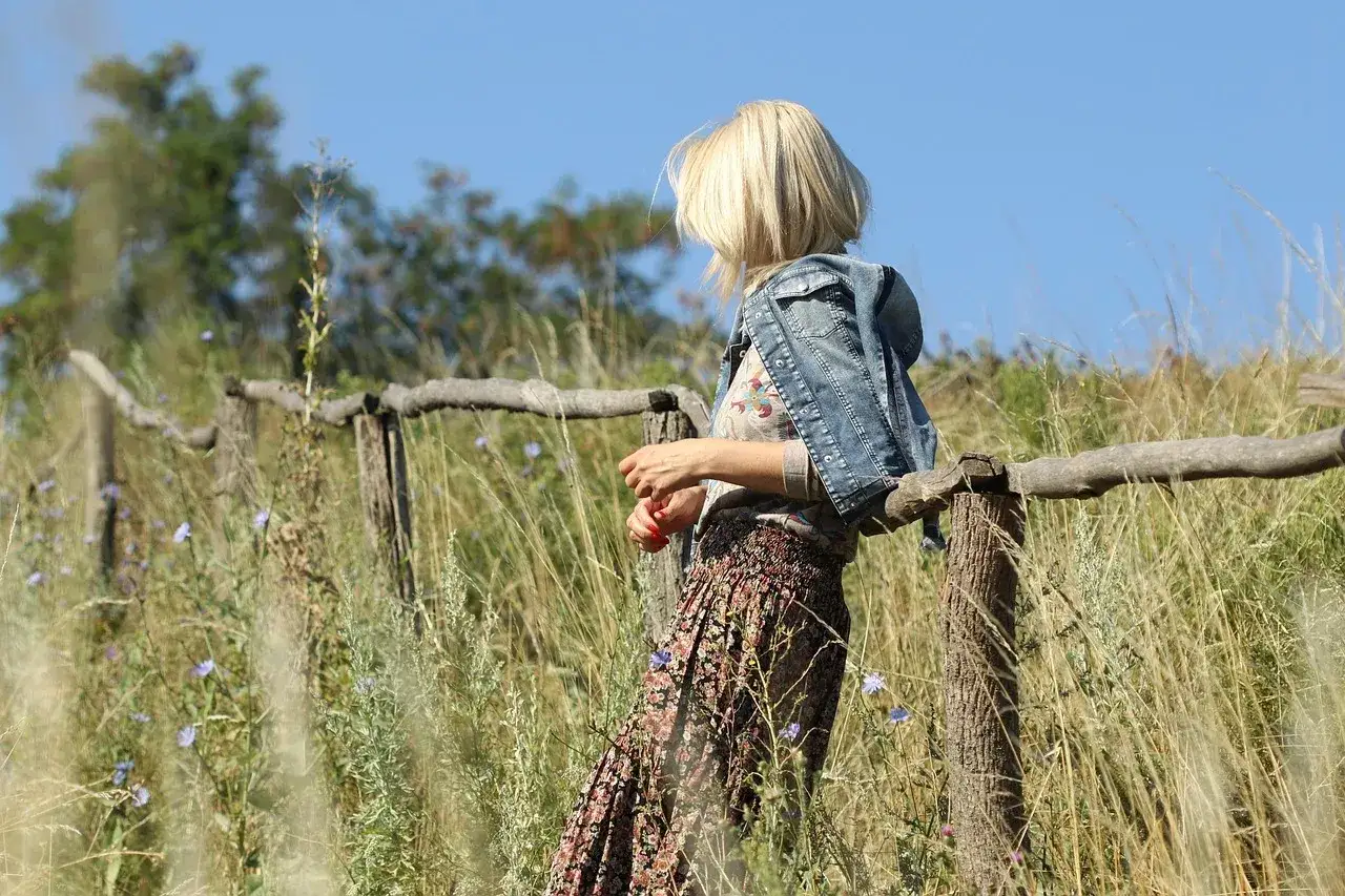 Blondynka w jeansowej kurtce i kwiecistej sp&oacute;dnicy, oparta o drewniane ogrodzenie. Styl boho, co jest teraz modne, idealnie wpisuje się w letni krajobraz.