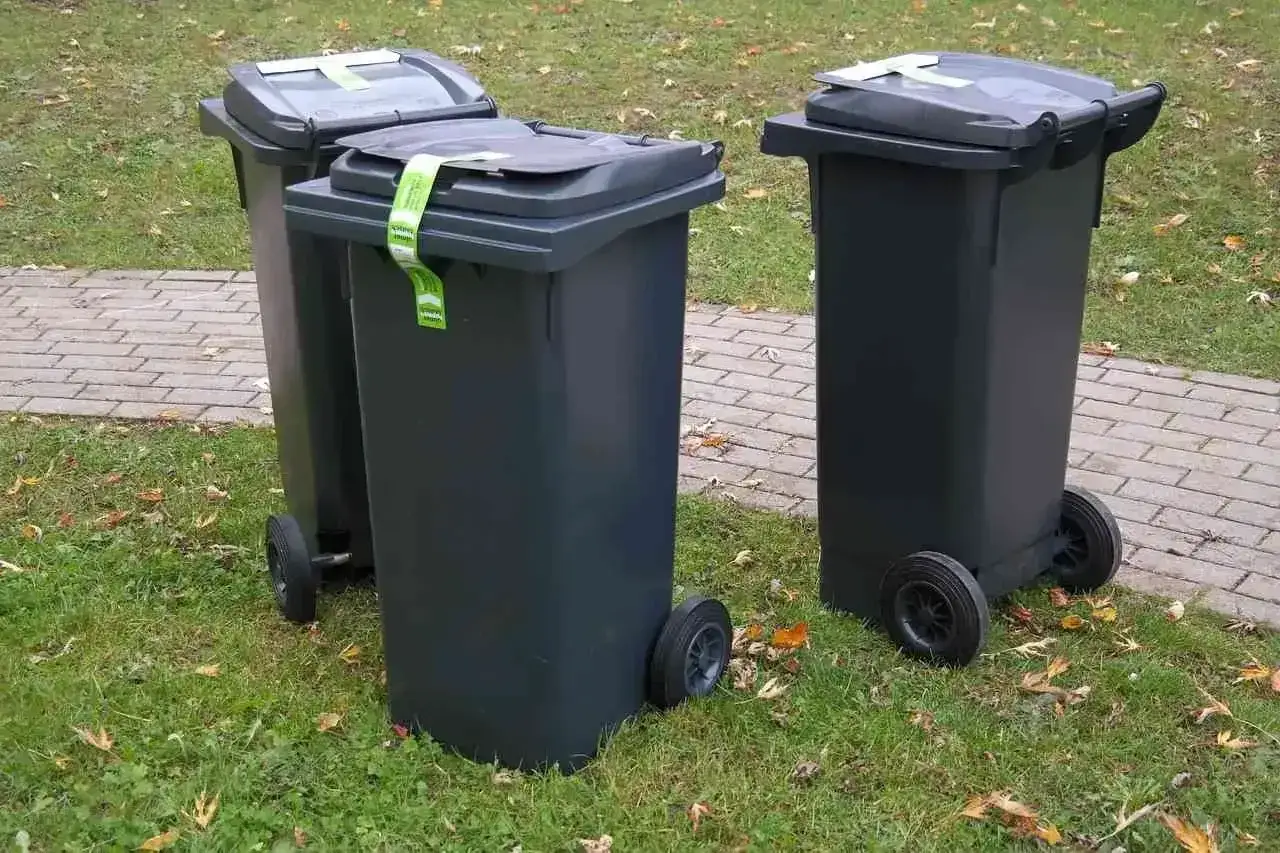 Trzy ciemnoszare kosze na śmieci stoją na trawniku obok chodnika. Jeden z nich ma zieloną naklejkę z napisem 