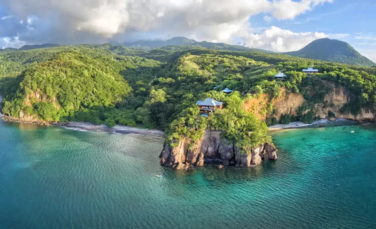 Vue aérienne de falaises verdoyantes sur l'île Dominique, avec des maisons perchées et une eau turquoise.