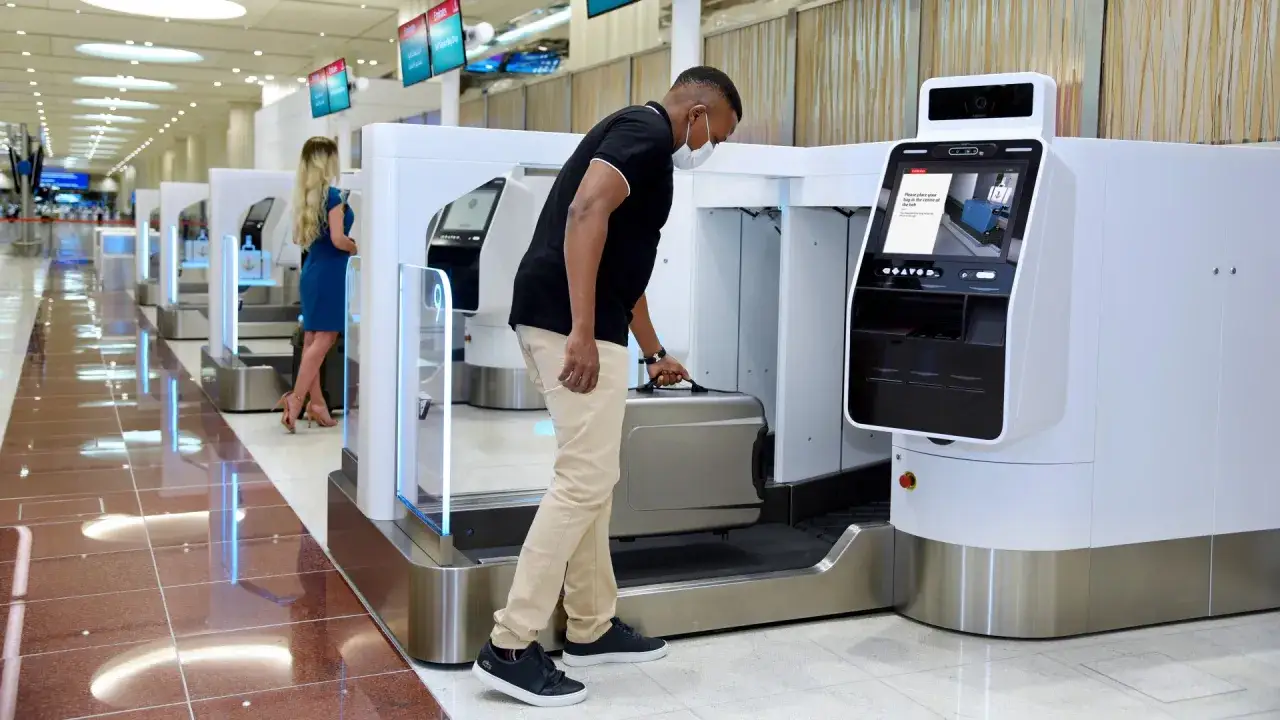 Mężczyzna w masce zakłada bagaż rejestrowany Emirates do samoobsługowej maszyny na lotnisku.