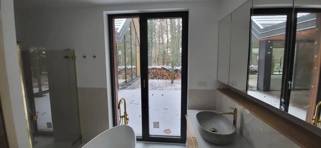 Nowoczesna łazienka z wanną i umywalką. Duże okna z czarnymi ramami, które podkreślają zmianę koloru okien plastikowych na bardziej elegancki.