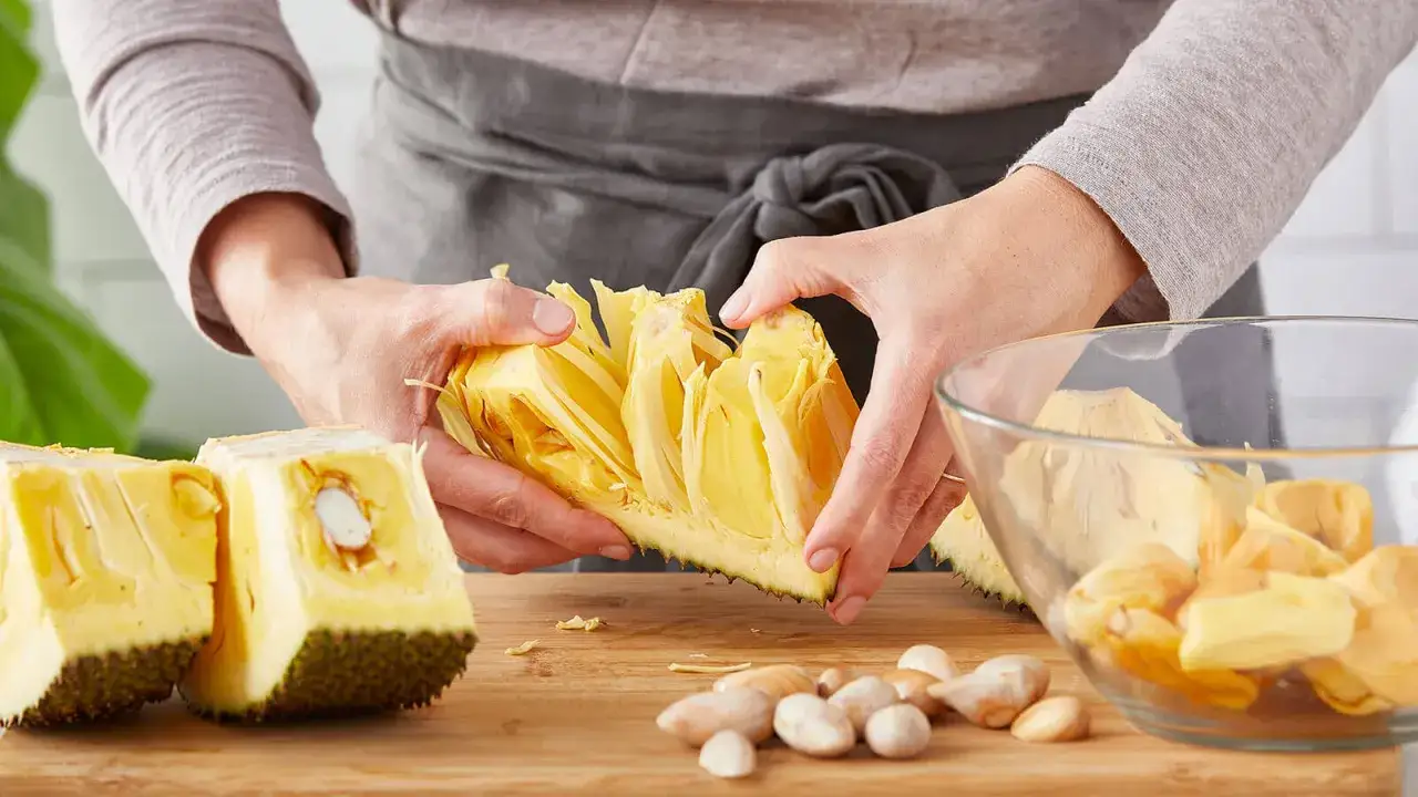 Odkryj, jackfruit co to jest! Ręce rozdzielają żółte, włókniste części owocu na desce. Obok leżą nasiona i miska z kawałkami jackfruita.