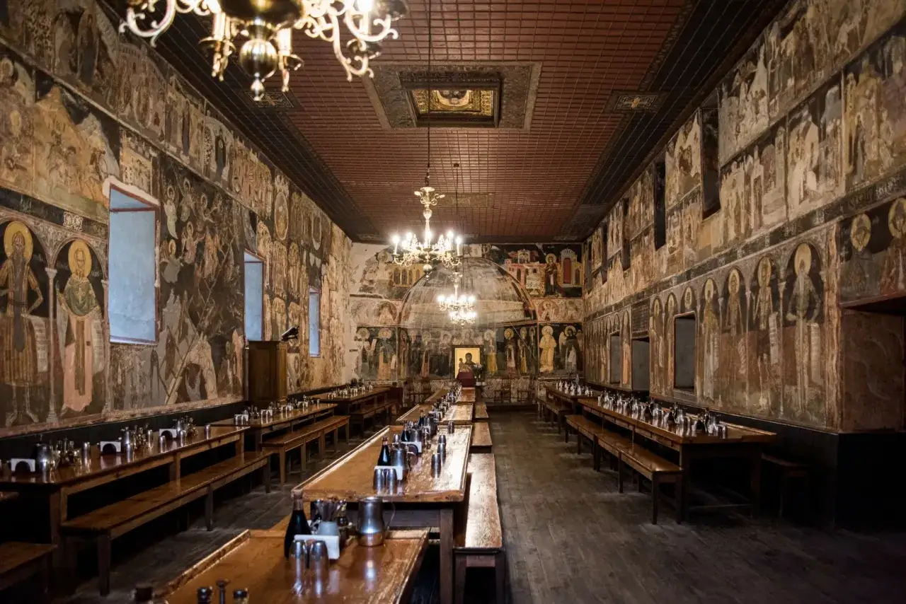 A refectory inside Mount Athos, with long wooden tables and benches, adorned with ancient frescoes depicting religious scenes.