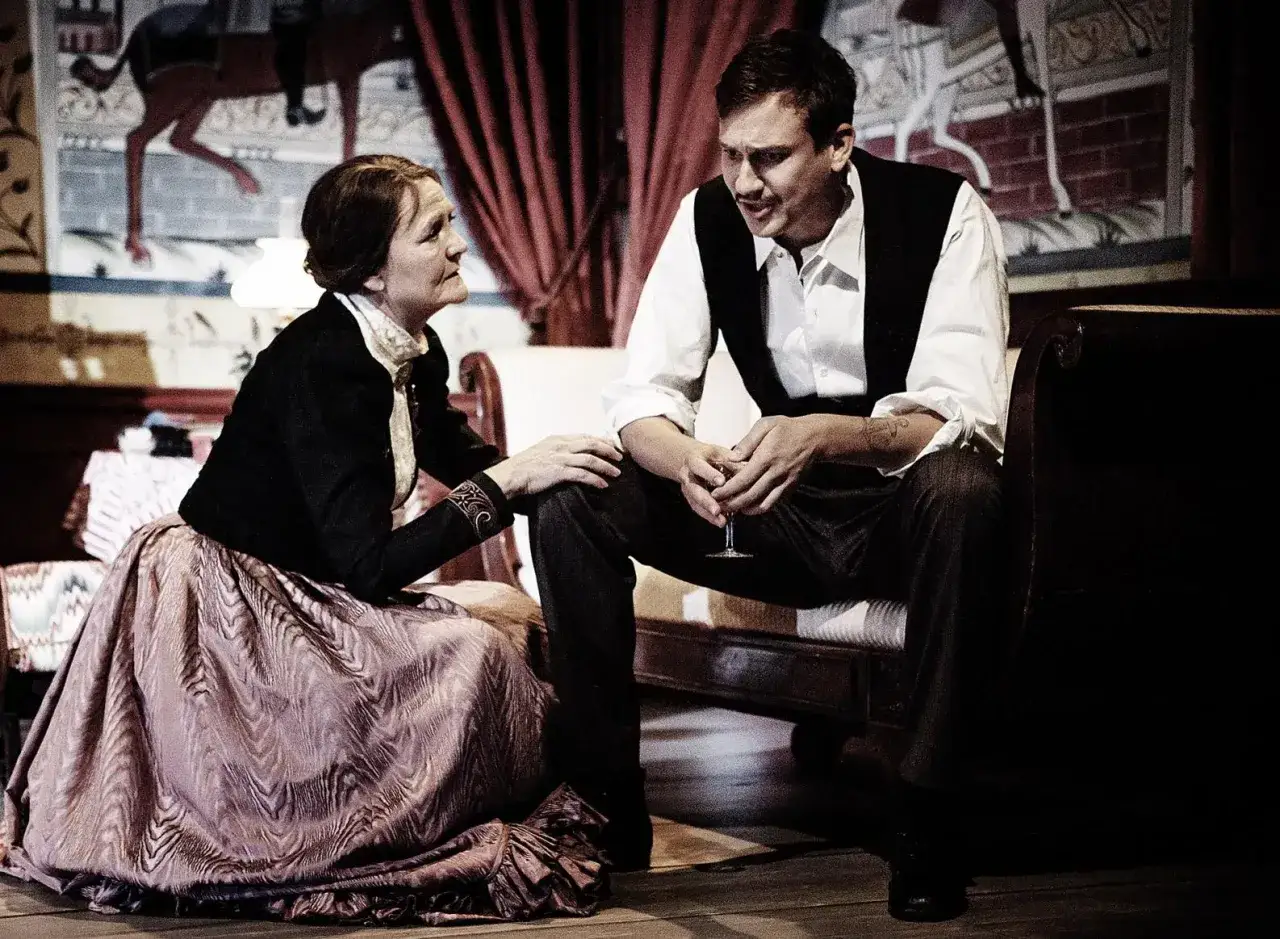 En kvinna i en stor kl&auml;nning och en man i v&auml;st sitter n&auml;ra varandra, i en scen som p&aring;minner om ett Ibsen-drama.