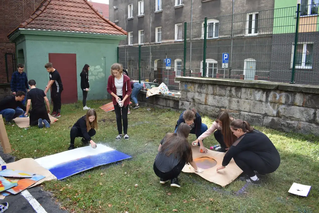 Młodzież tworzy sztukę na trawie, jakby lekcja plastyki w liceum przeniosła się na zewnątrz.