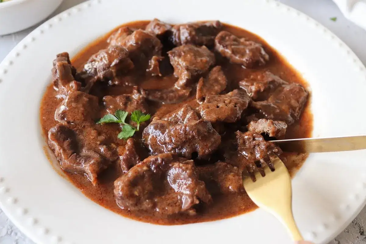 Plato de morcillo de ternera estofado en salsa, servido en una fuente blanca. Un tenedor dorado corta un trozo tierno.