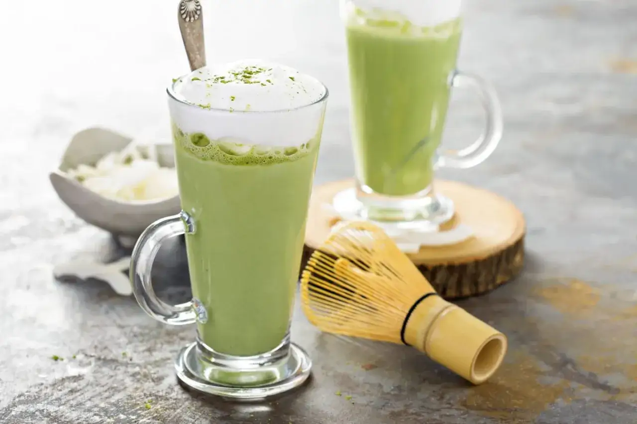 Zielony, spieniony nap&oacute;j w szklance, czyli matcha latte. Obok bambusowa miotełka do ubijania.