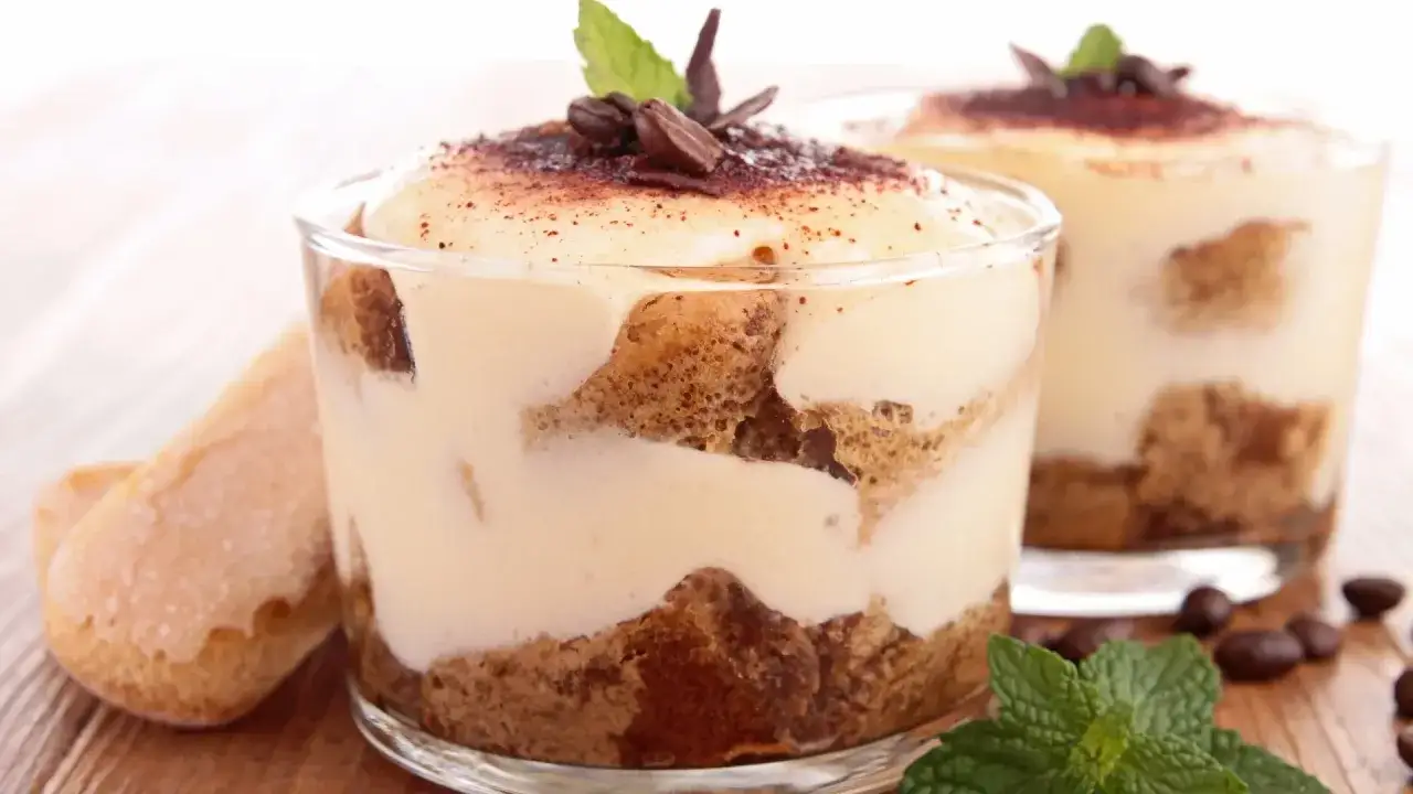 Zwei Gläser mit cremigem Tiramisu originale, garniert mit Kakaopulver, Kaffeebohnen und Minze. Löffelbiskuits liegen daneben.