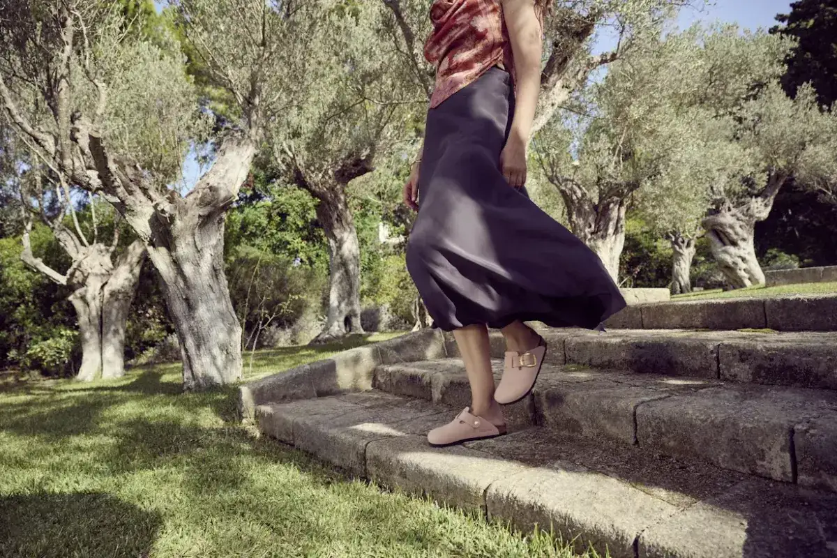 Kobieta w zwiewnej sukience schodzi po kamiennych schodach w różowych klapkach, które przypominają Bayton lub Birkenstock.