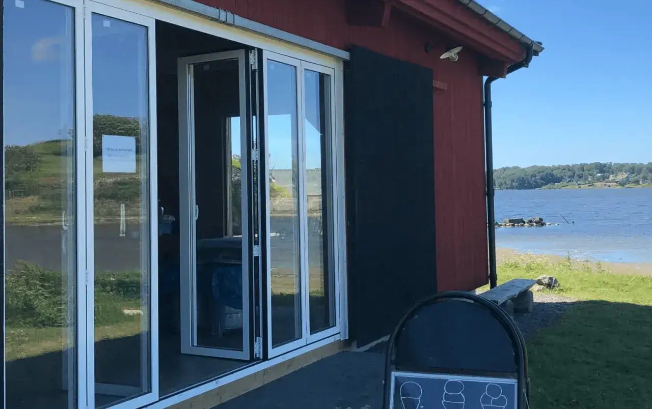 En röd byggnad vid vattnet med stora glasdörrar. Perfekt för en fika ute med utsikt över sjön.