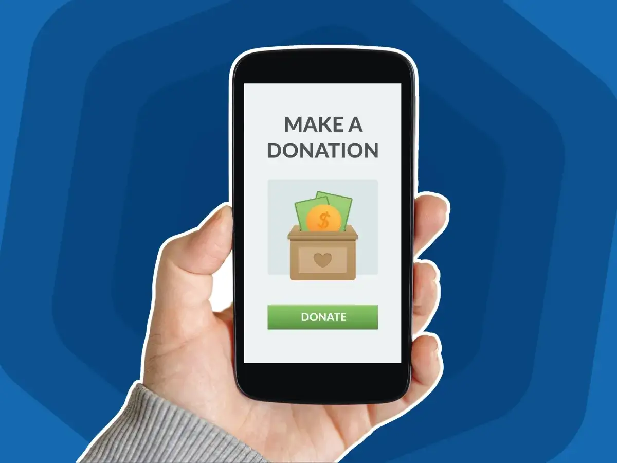 A hand holds a smartphone displaying a donation screen. The screen shows a coin and bills going into a donation box, with a heart symbol. The text 