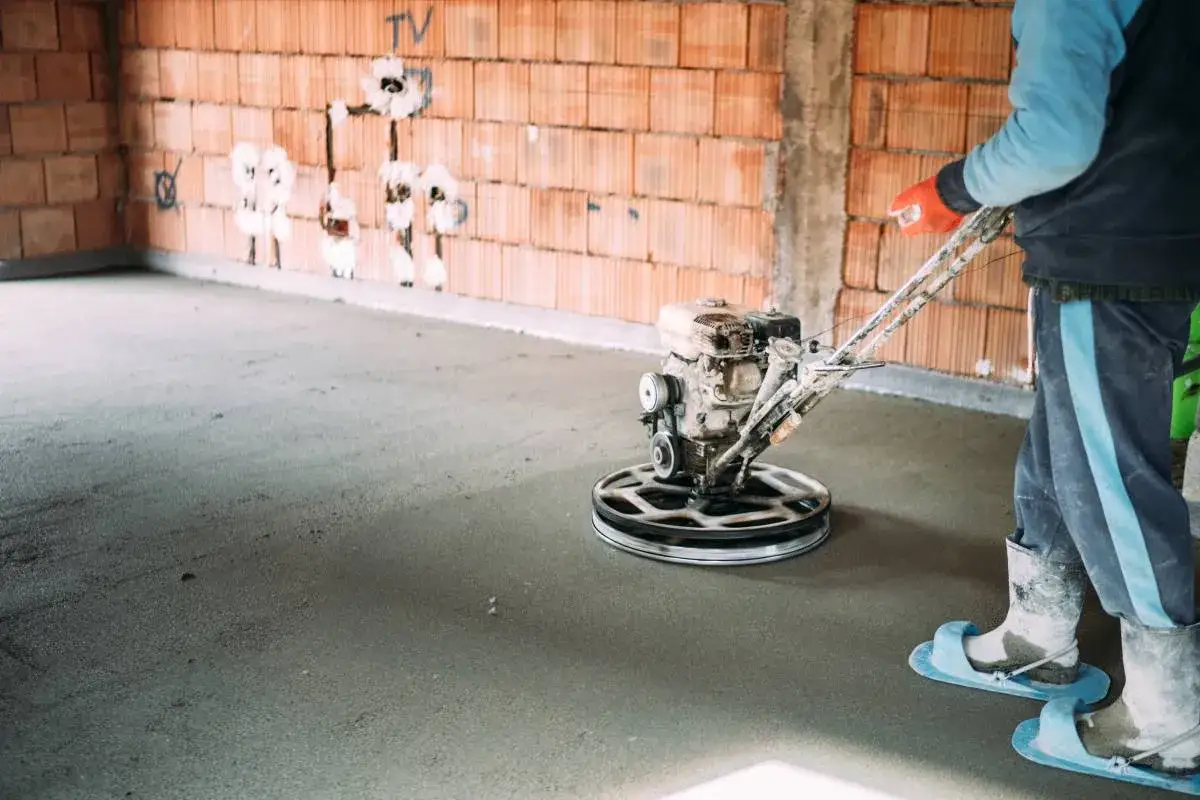 Pracownik wykonuje wylewanie posadzki za pomocą zacieraczki mechanicznej. Widać ceglane ściany i świeży beton.
