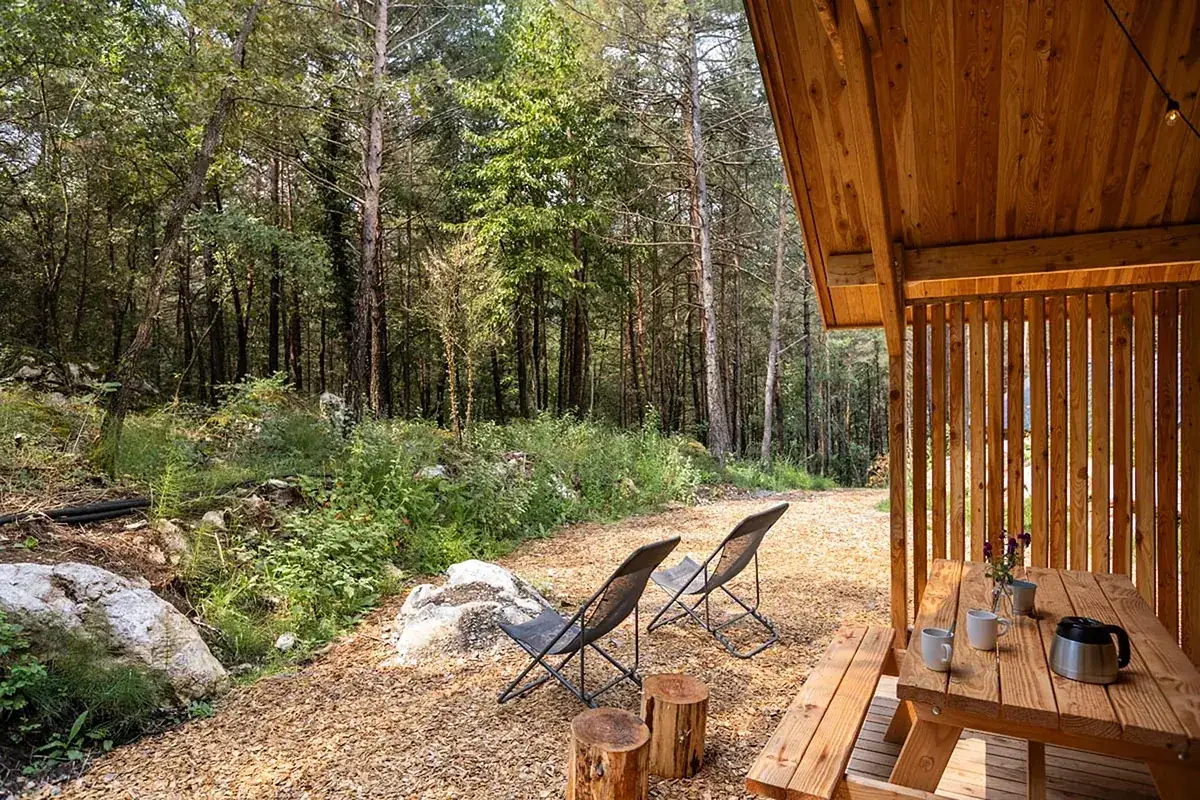 Dos sillas plegables y mesa de picnic bajo un porche de madera, rodeados de bosque. Un lugar idílico para disfrutar de los mejores camping de España.