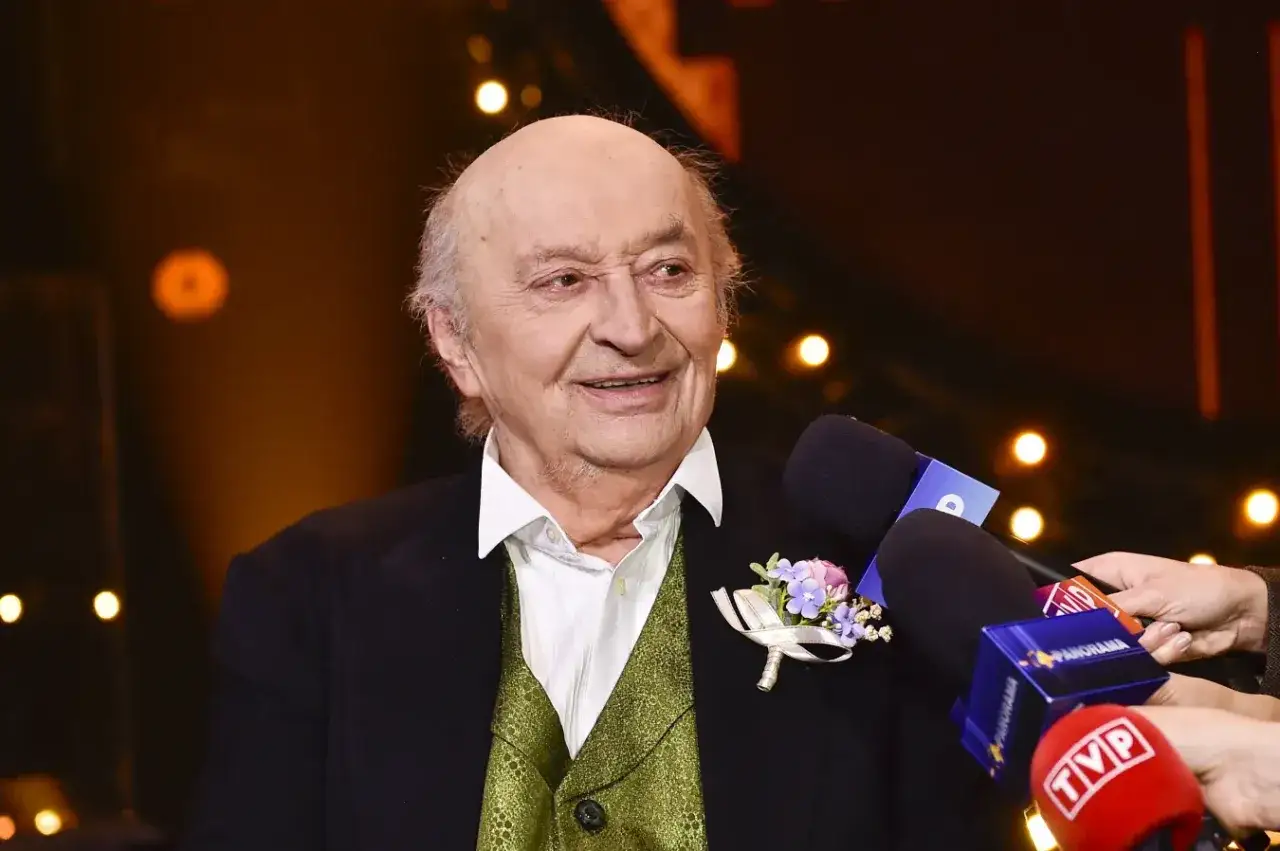 Uśmiechnięty Bohdan Łazuka w eleganckim stroju, otoczony przez mikrofony mediów, udziela wywiadu.