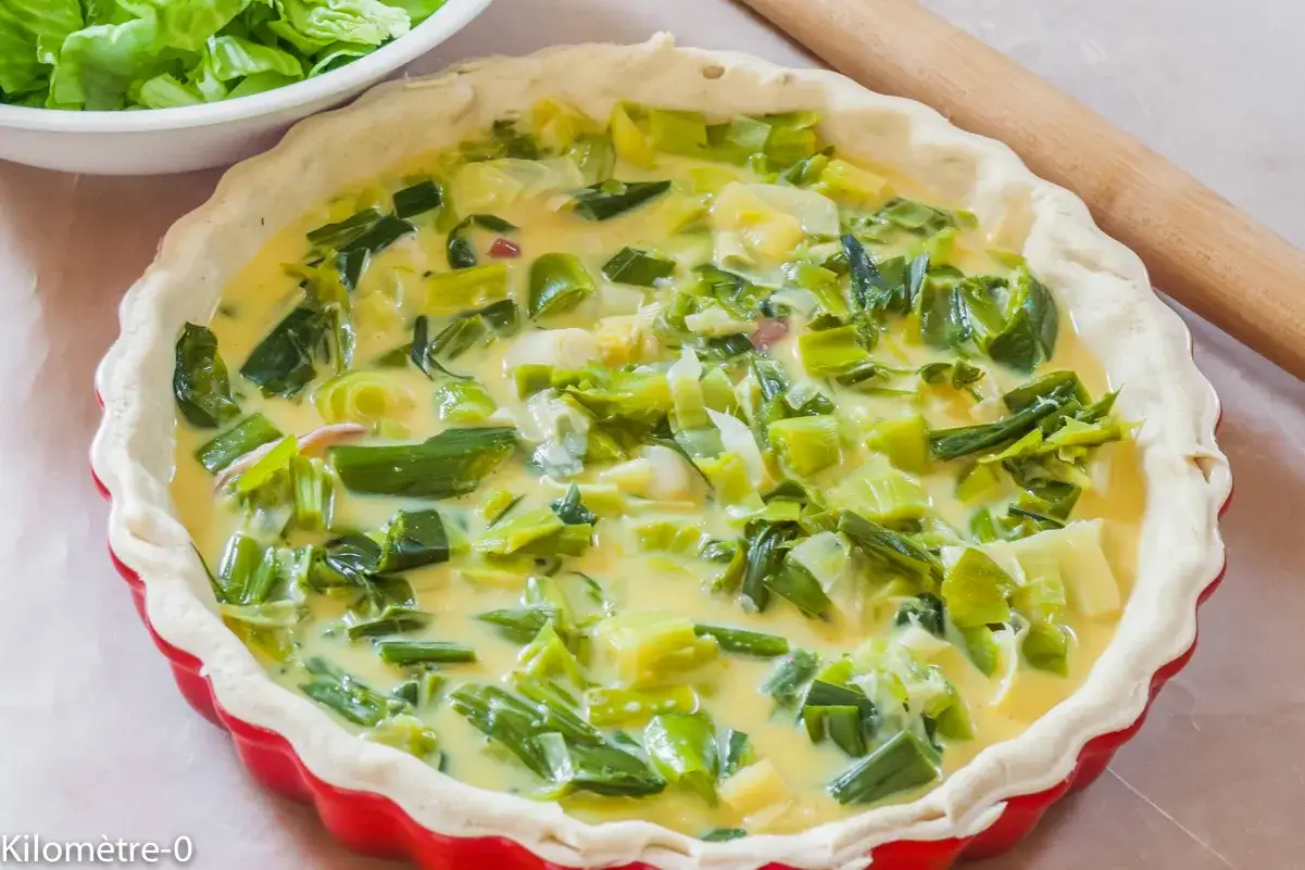 Tarte aux poireaux crue dans un moule rouge, prête à cuire. Un accompagnement parfait pour un repas léger.