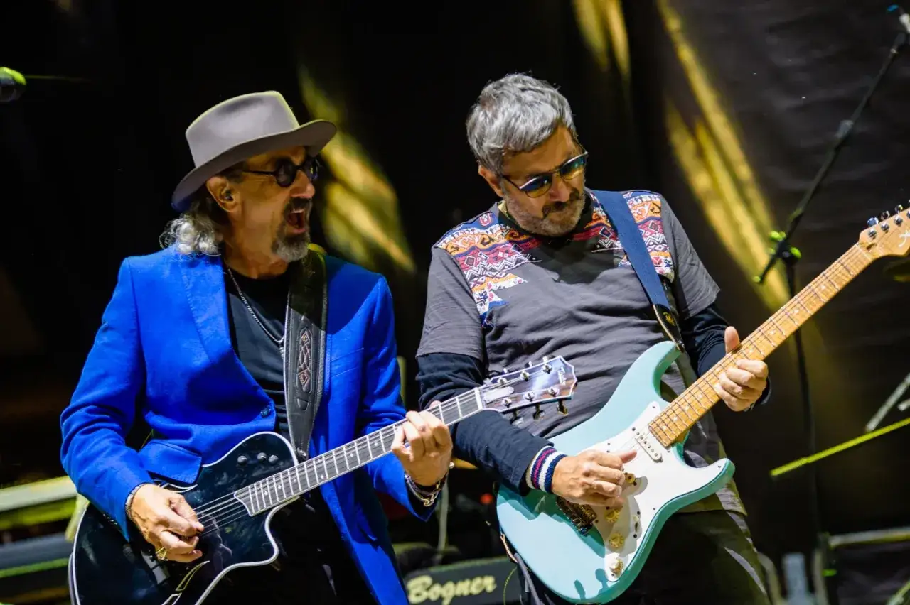 Dwóch gitarzystów na scenie, jeden w niebieskiej marynarce i kapeluszu, drugi w koszulce z etnicznym wzorem. Ich gra to prawdziwy **dire straits legacy**.