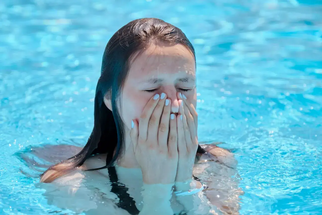 A girl in a pool covers her nose and mouth, eyes closed, possibly experiencing a chlorine allergy sneezing fit.