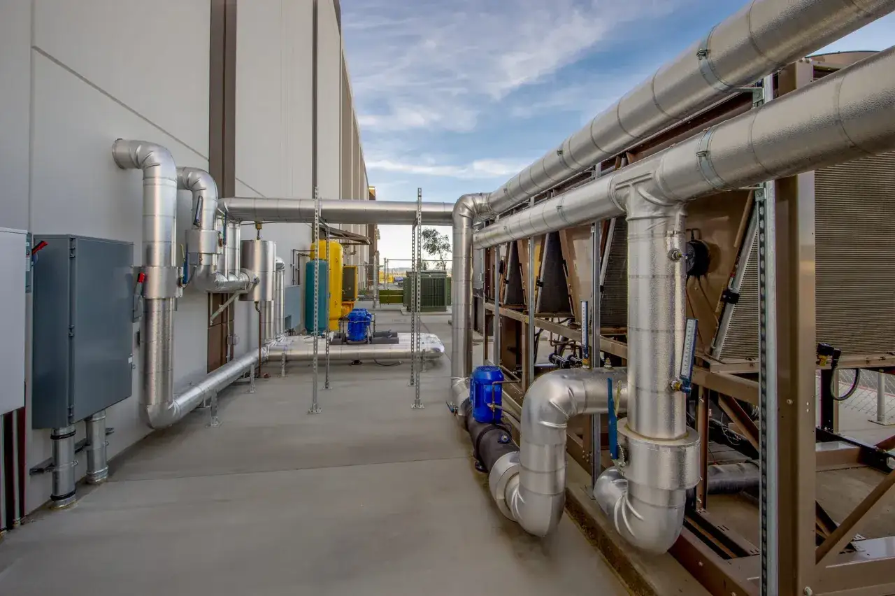 Instalación industrial con grandes tuberías plateadas, parte de sistemas de ventilación y extracción de aire, bajo un cielo azul con nubes.