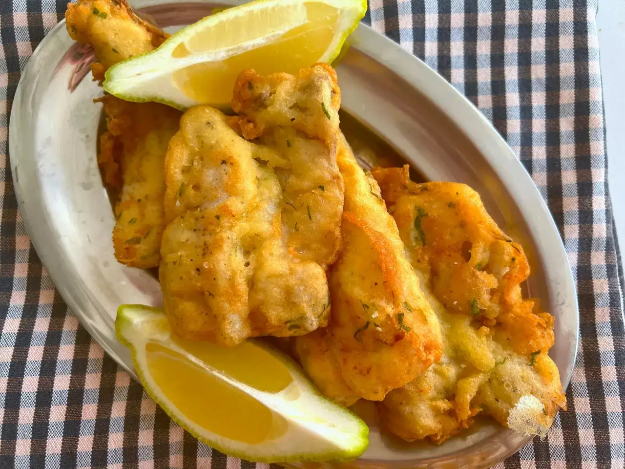 Crujiente merluza a la romana, dorada y servida con gajos de lim&oacute;n fresco en una bandeja met&aacute;lica sobre un mantel a cuadros.