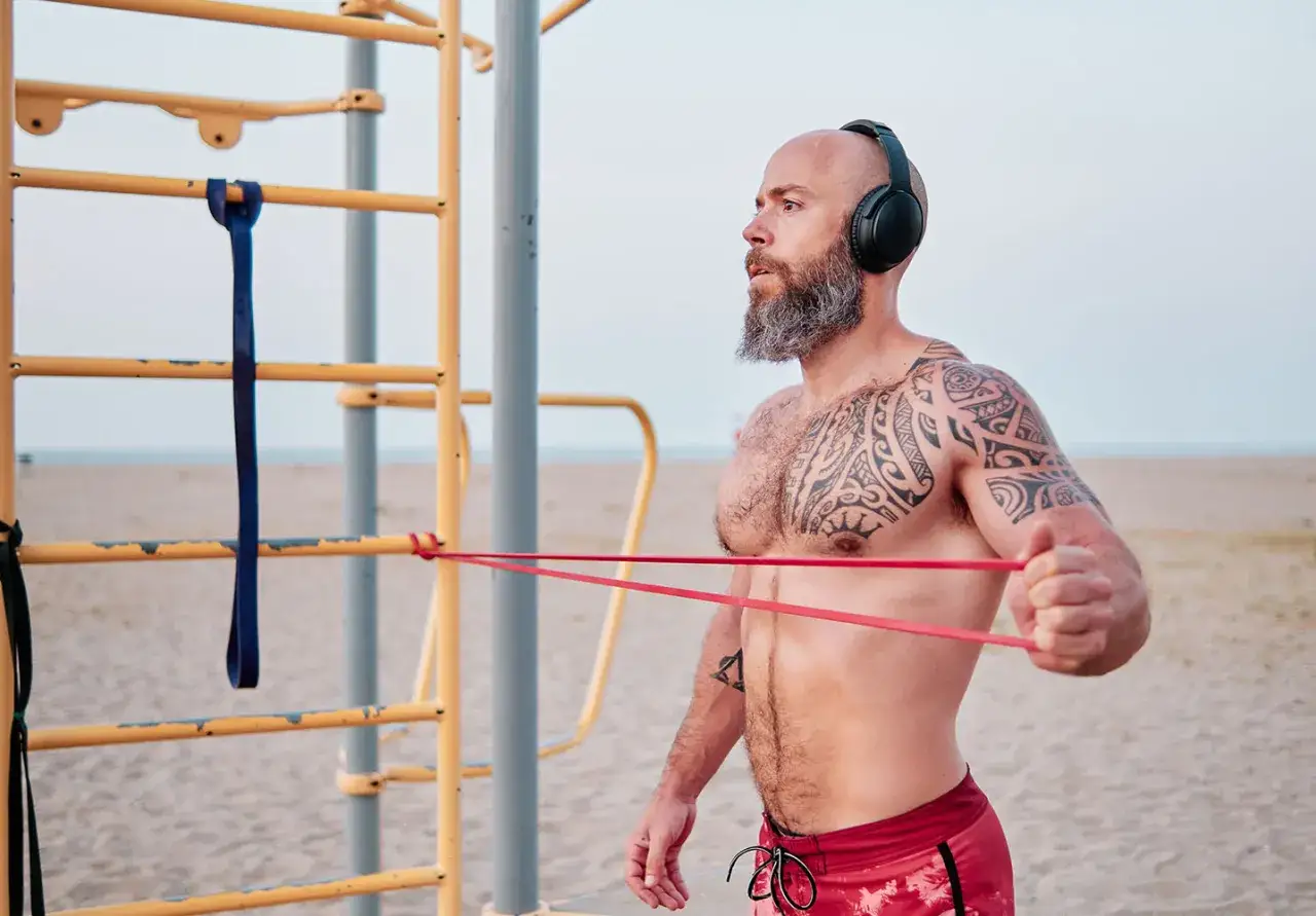 Man med tatueringar och skägg tränar med gummisnoddar vid ett utegym på stranden.